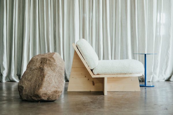 Side view of a wooden-framed upholstered sofa with a blue side table – stone detail on the left, white curtain in the background.