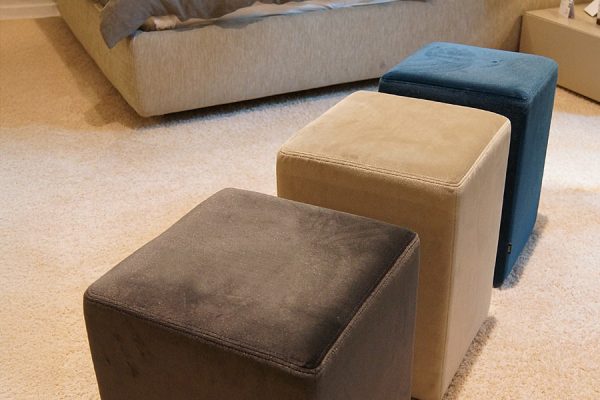Three differently colored POMP stools placed in a cozy bedroom next to a bed.