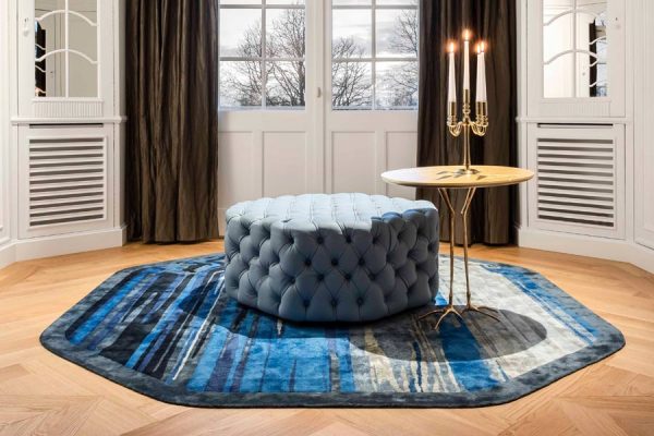 Interior with hexagonal designer carpet in blue and grey tones, paired with a tufted ottoman and brass table – from the 'Daydreams' collection by Olaf Kitzig.