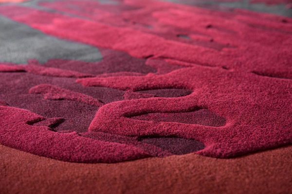 Close-up of textured carpet surface in rich red hues with abstract relief patterns – emphasizing material depth and color layering.