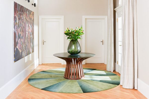 Bright hallway with round green and blue carpet under a sculptural wooden table with flowers.